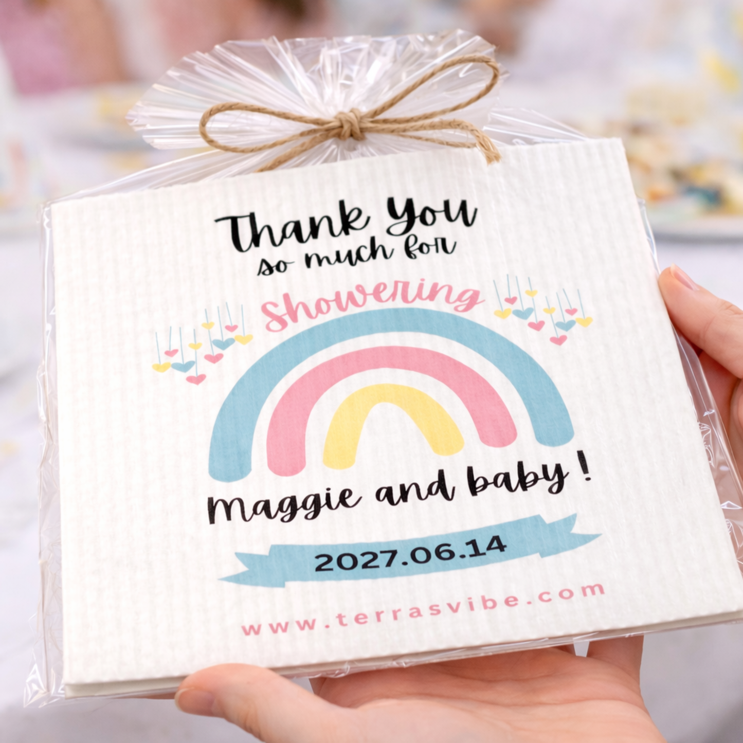 Pack of swedish cloth baby shower favors with a rainbow design and personalized text, held by a hand.