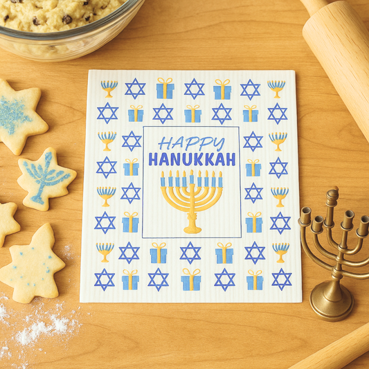 Happy Hanukkah swedish  dishcloth with menorah and star designs on a wooden table with cookies and a candle.
