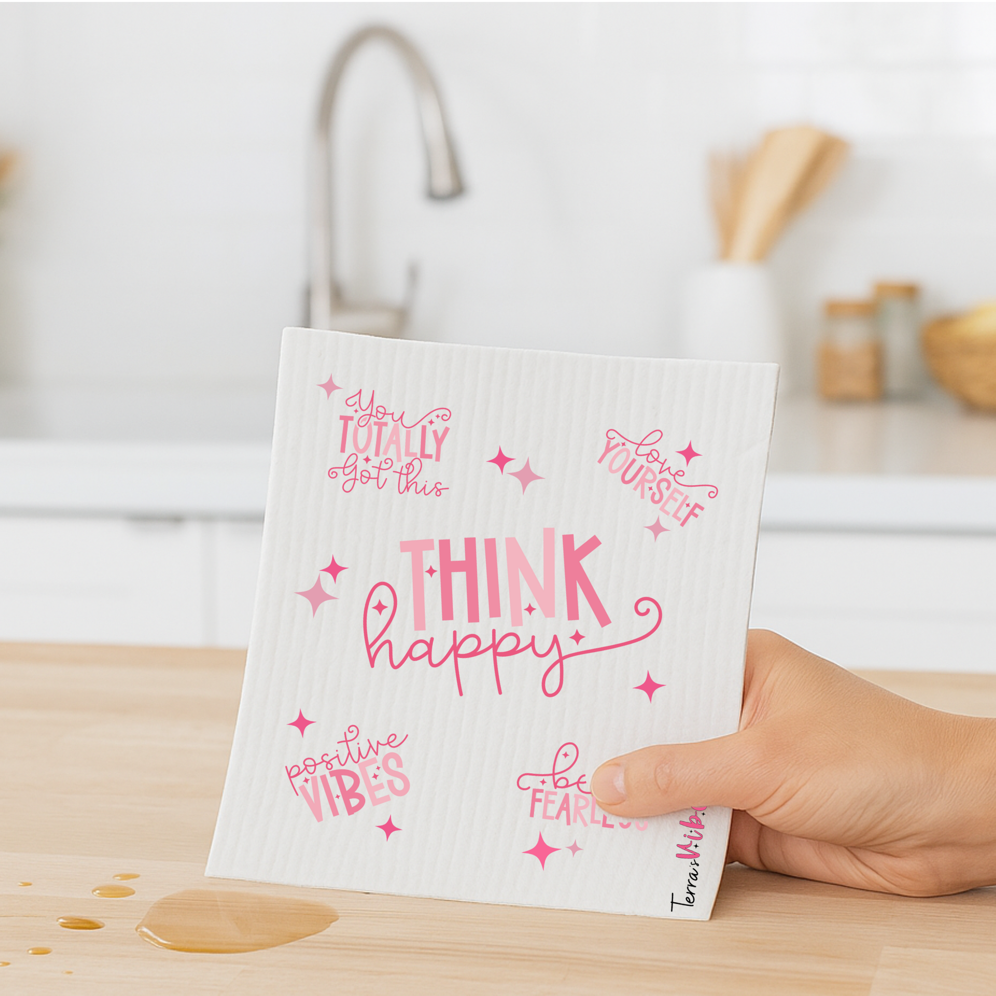 Hand holding a white swedish dishcloth with pink text and designs on a kitchen counter.